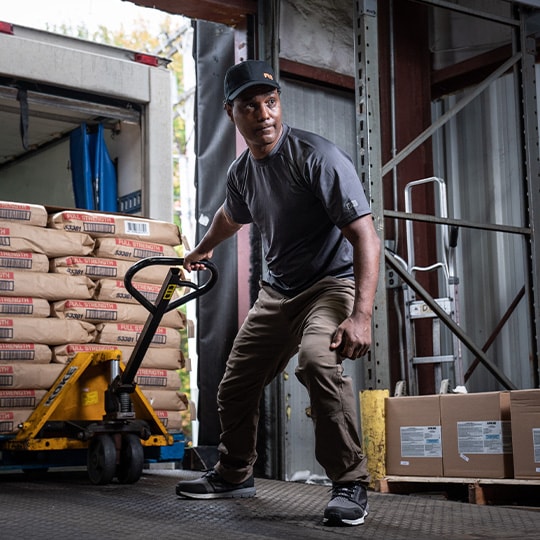 Image of a man in a warehouse, wearing dark Timberland work clothing and black work sneakers, pulling a pallet of concrete.