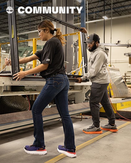 Image of a man and woman on an assembly line, carrying a black frame of a window. They are both wearing safety goggles, Timberland work shirts and pants, and athletic Timberland work shoes with large outsoles.